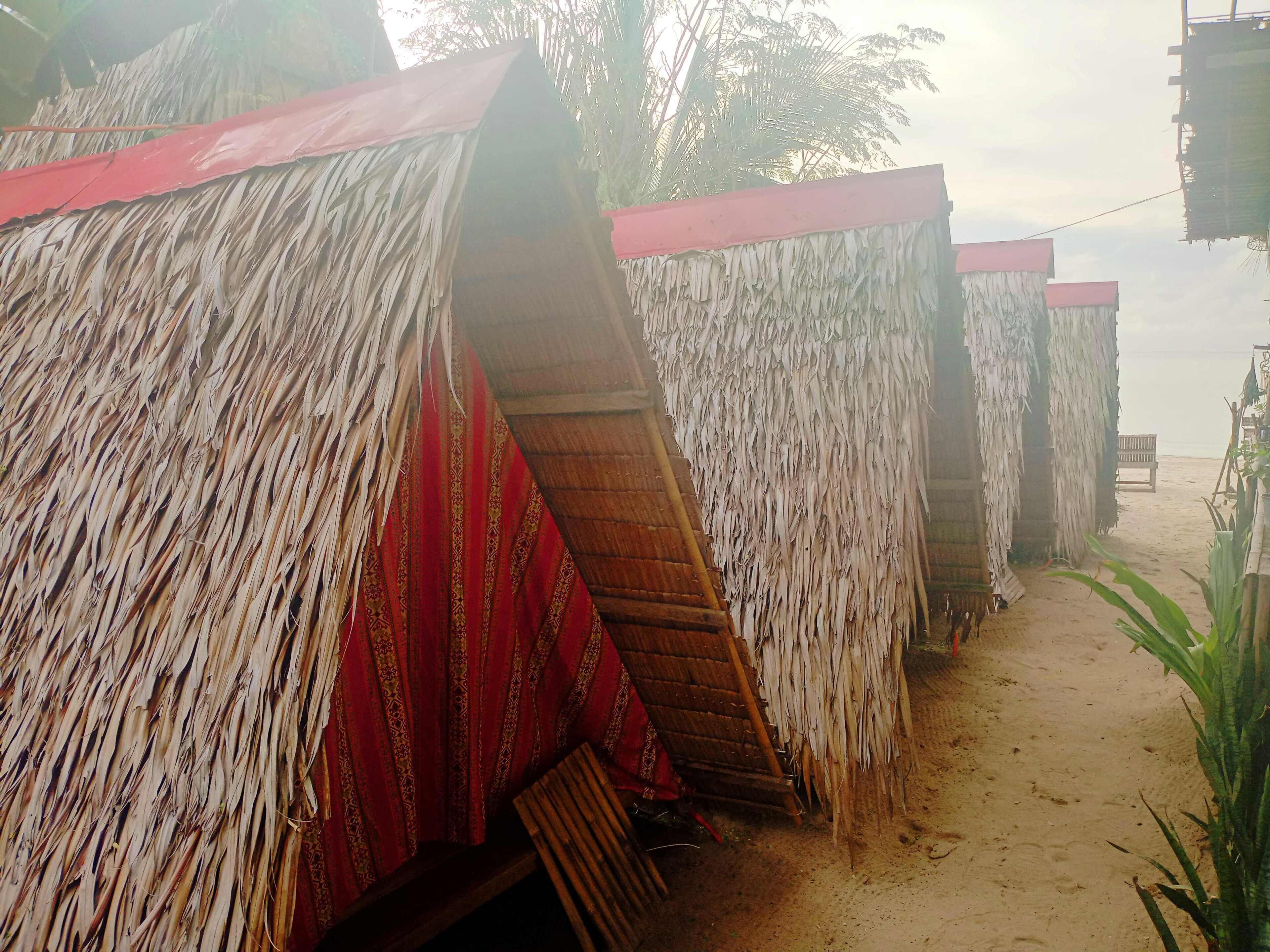 A row of thatched-roof huts with red and white striped awnings, wooden benches, and a sandy path leading to a beach.