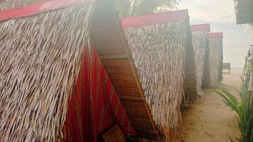 A row of thatched-roof huts with red and white striped awnings, wooden benches, and a sandy path leading to a beach.