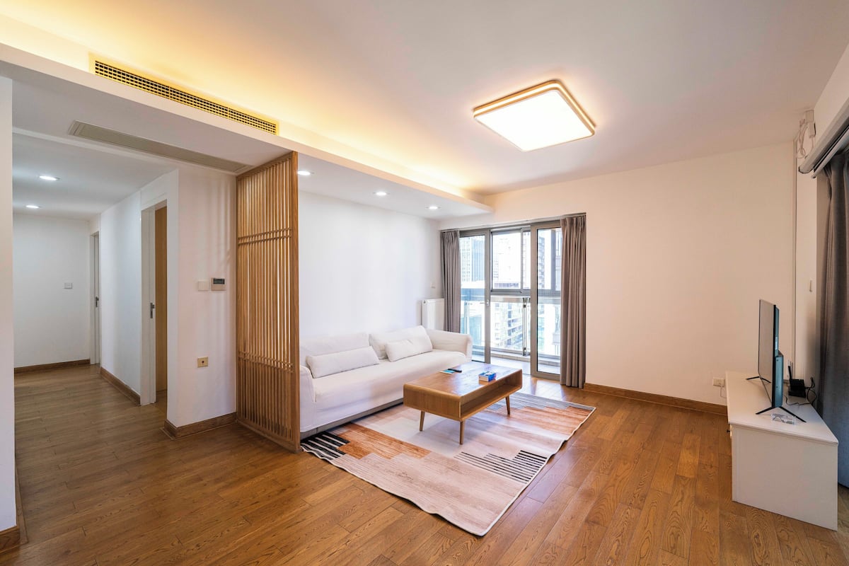 A modern living room with a wooden floor, a white sofa, a coffee table, and a large window with curtains.