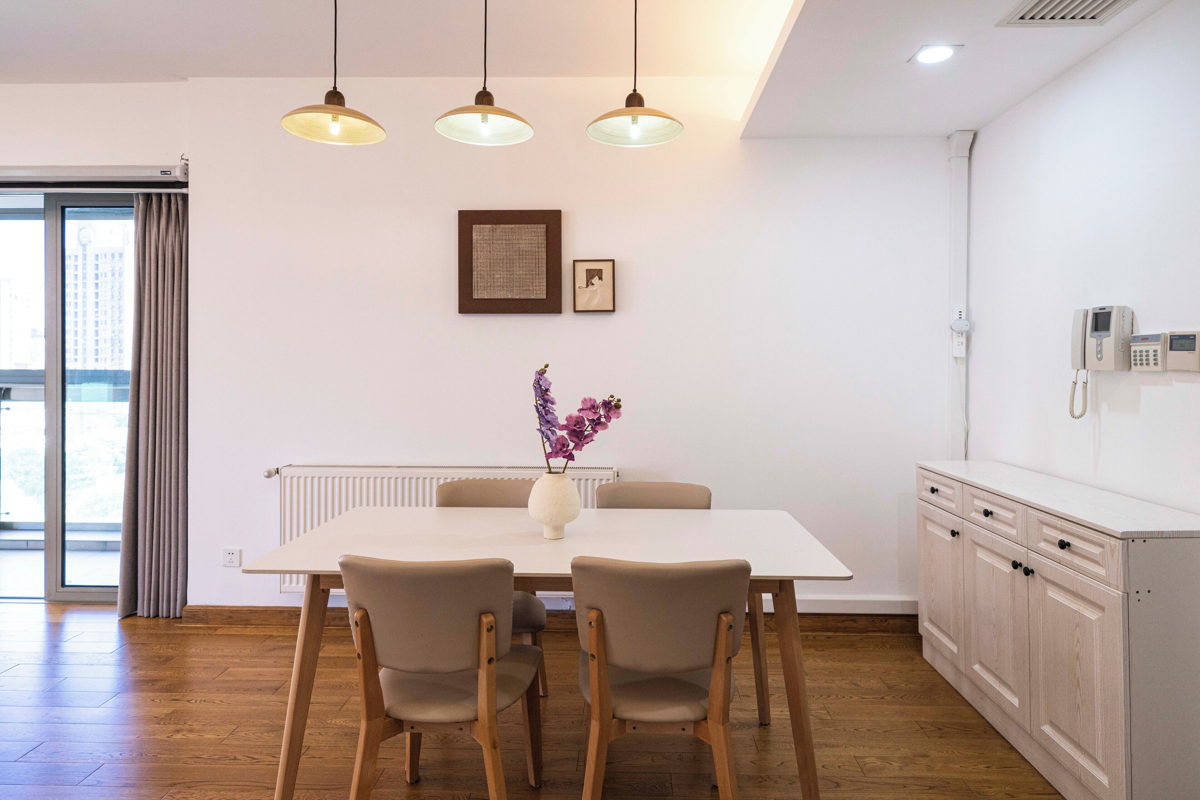 A modern dining area with a white table and four chairs, a vase with flowers, pendant lights, and a white cabinet with a phone.