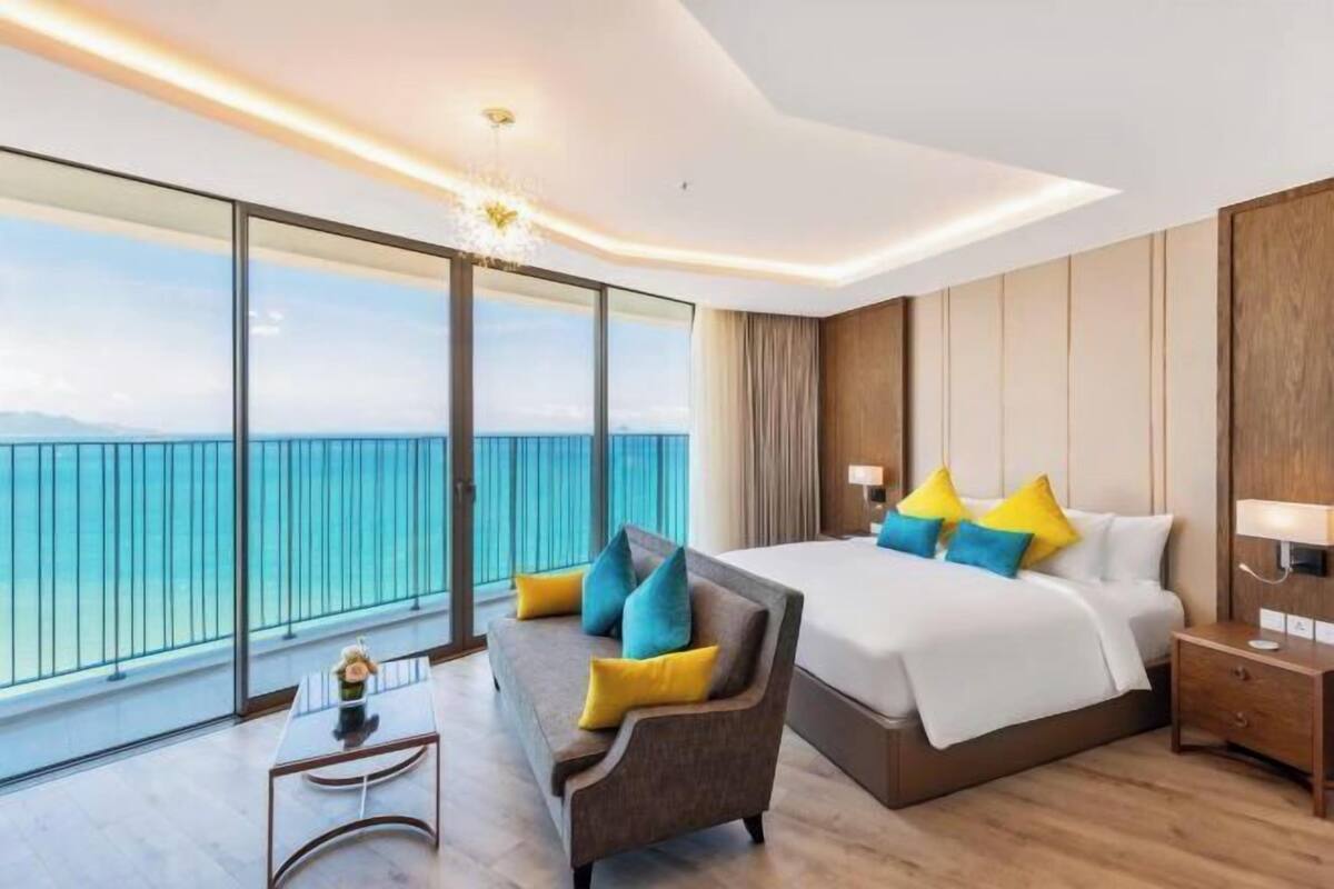 Ein modernes Hotelzimmer mit einem großen Bett, einer Couch, einem kleinen Tisch und einem Nachttisch. Das Zimmer verfügt über große Fenster mit Blick aufs Meer.