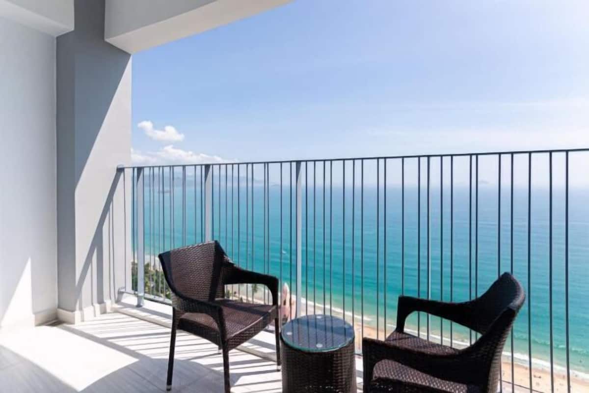 Ein Balkon mit zwei Korbstühlen und einem kleinen runden Tisch, mit Blick auf einen Strand und klares blaues Wasser.