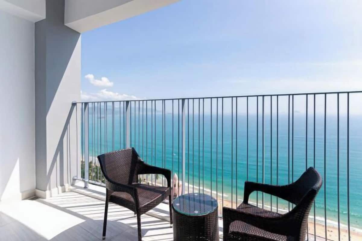 Ein Balkon mit zwei Korbstühlen und einem kleinen runden Tisch mit Blick auf einen Strand und den Ozean.