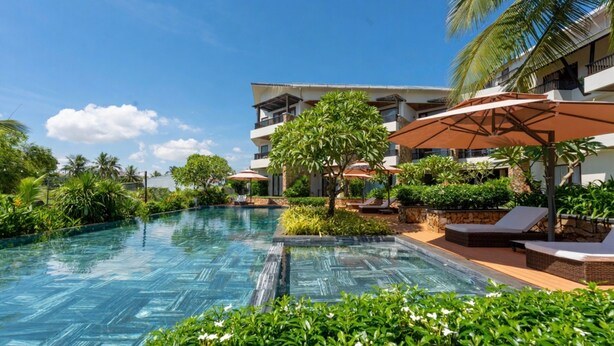 A swimming pool with lounge chairs and umbrellas, surrounded by greenery and a building with balconies. - Hoi An Eco Retreat (Da Nang)