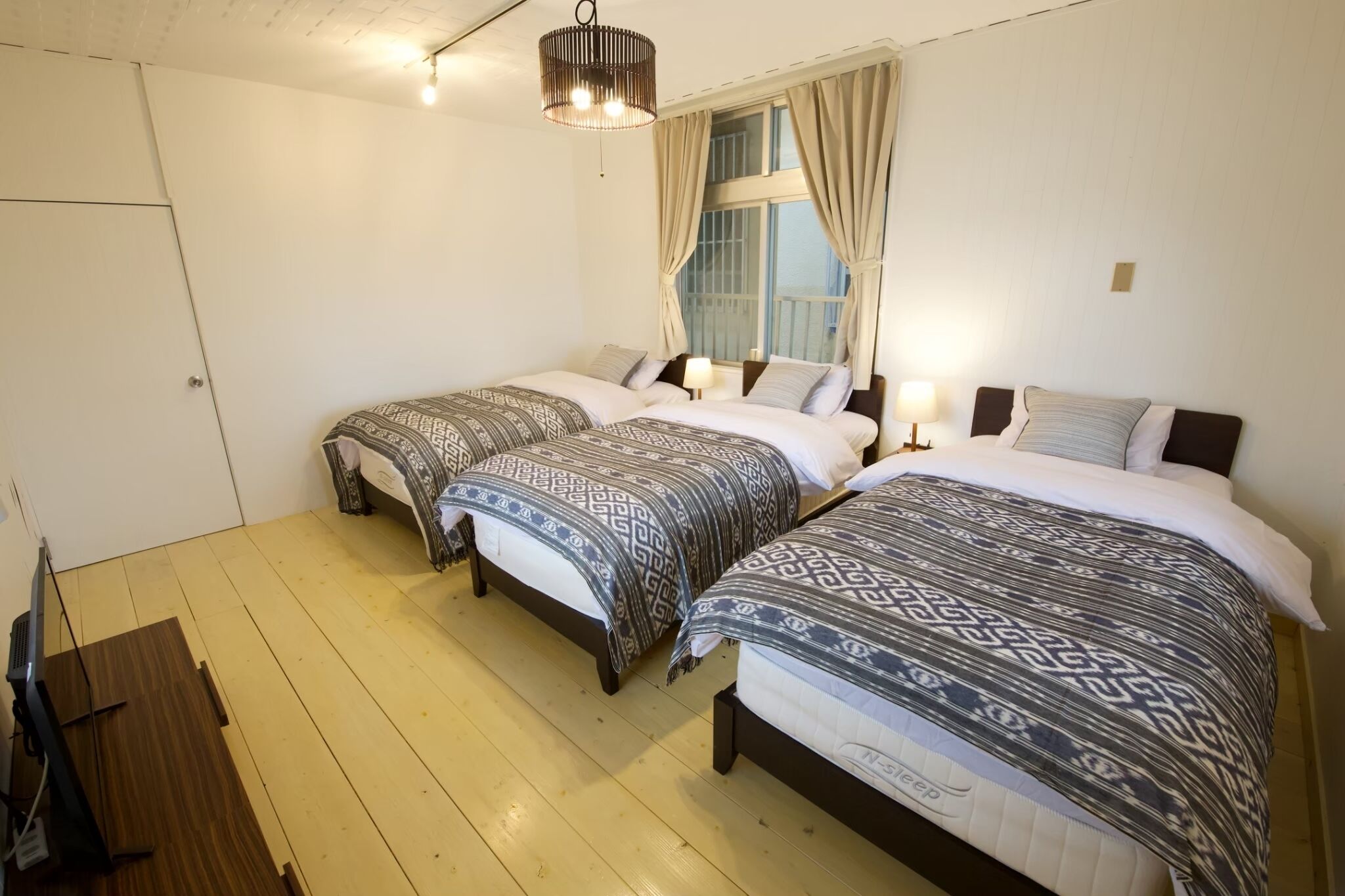 Une chambre avec trois lits, chacun agrémenté de draps à motifs, un plancher en bois, un luminaire au plafond et une fenêtre avec des rideaux.