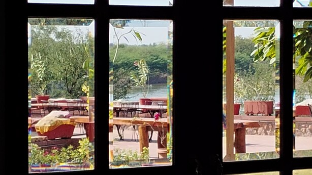 Una vista attraverso una finestra a più vetri, che mostra una zona con posti a sedere all'aperto, tavoli e sedie, un prato e un albero.