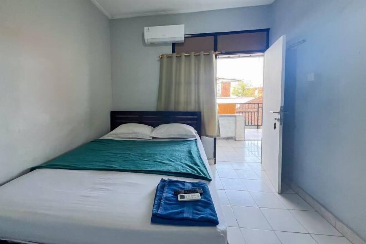 A single bed with a green blanket, a white pillow, and a remote control on the bed. There is a window with beige curtains, an air conditioning unit on the wall, and a door leading to an outdoor area.