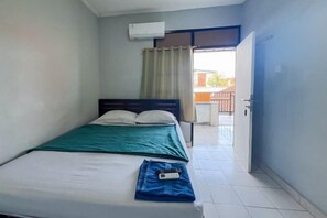 A single bed with a green blanket, a white pillow, and a remote control on the bed. There is a window with beige curtains, an air conditioning unit on the wall, and a door leading to an outdoor area.