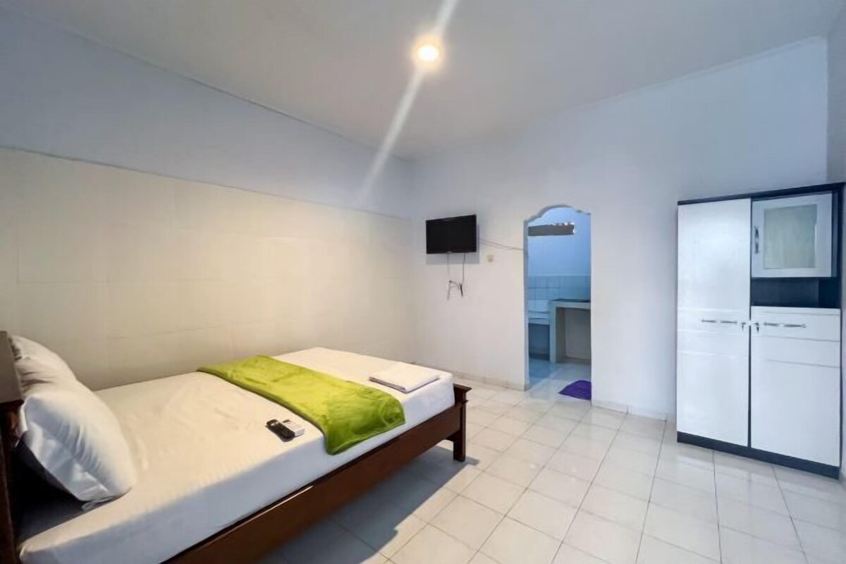 A single bed with a green blanket, a white pillow, a mounted TV, and a wardrobe in a room with white walls and tiled flooring.