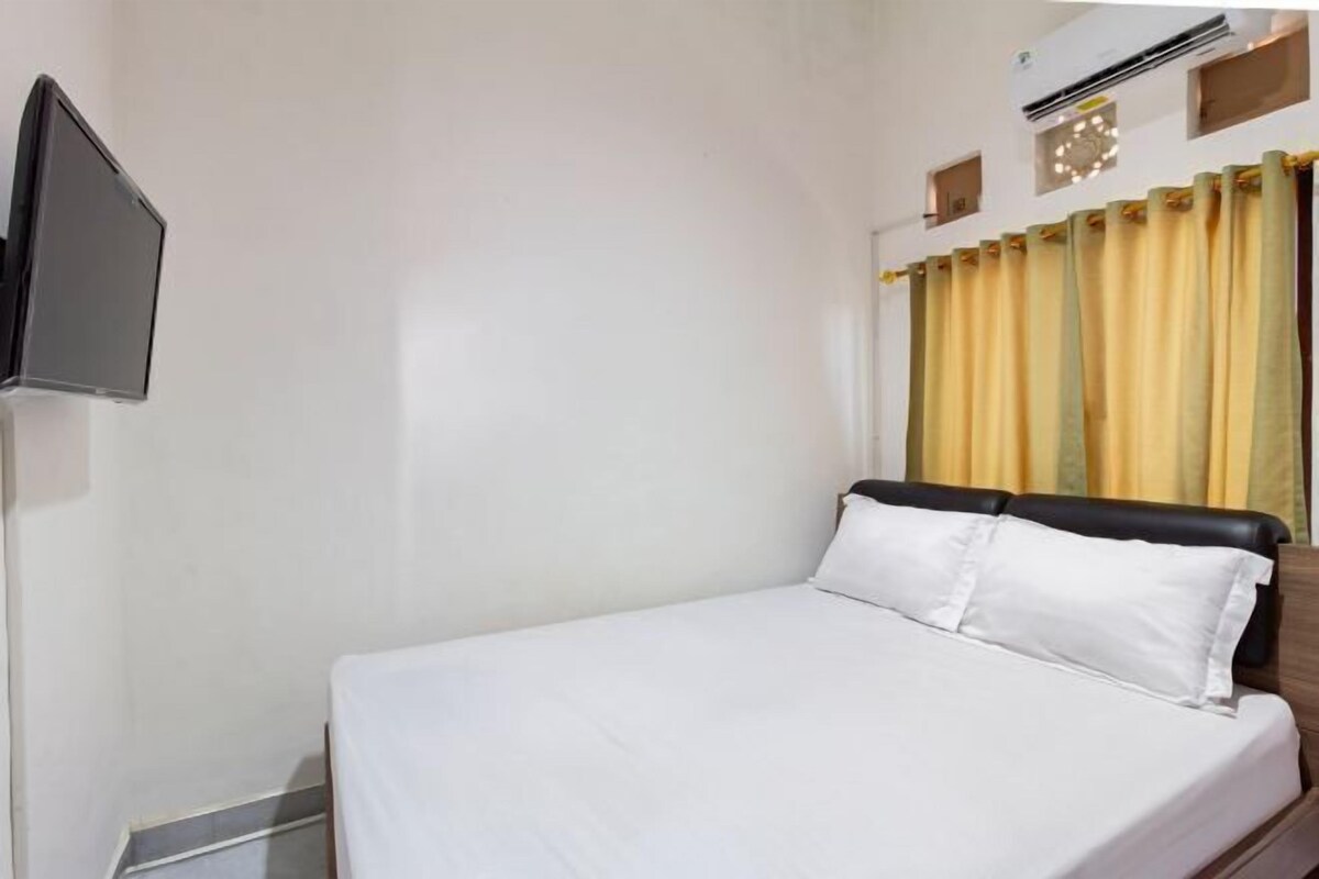 A single bed with white bedding, a television mounted on the wall, and a window with yellow curtains.