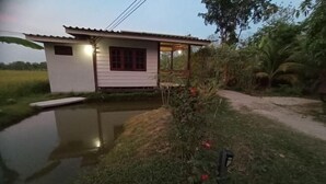 A small, single-story house with a porch, surrounded by a pond and lush vegetation. - ID Garden Home (Lampang)