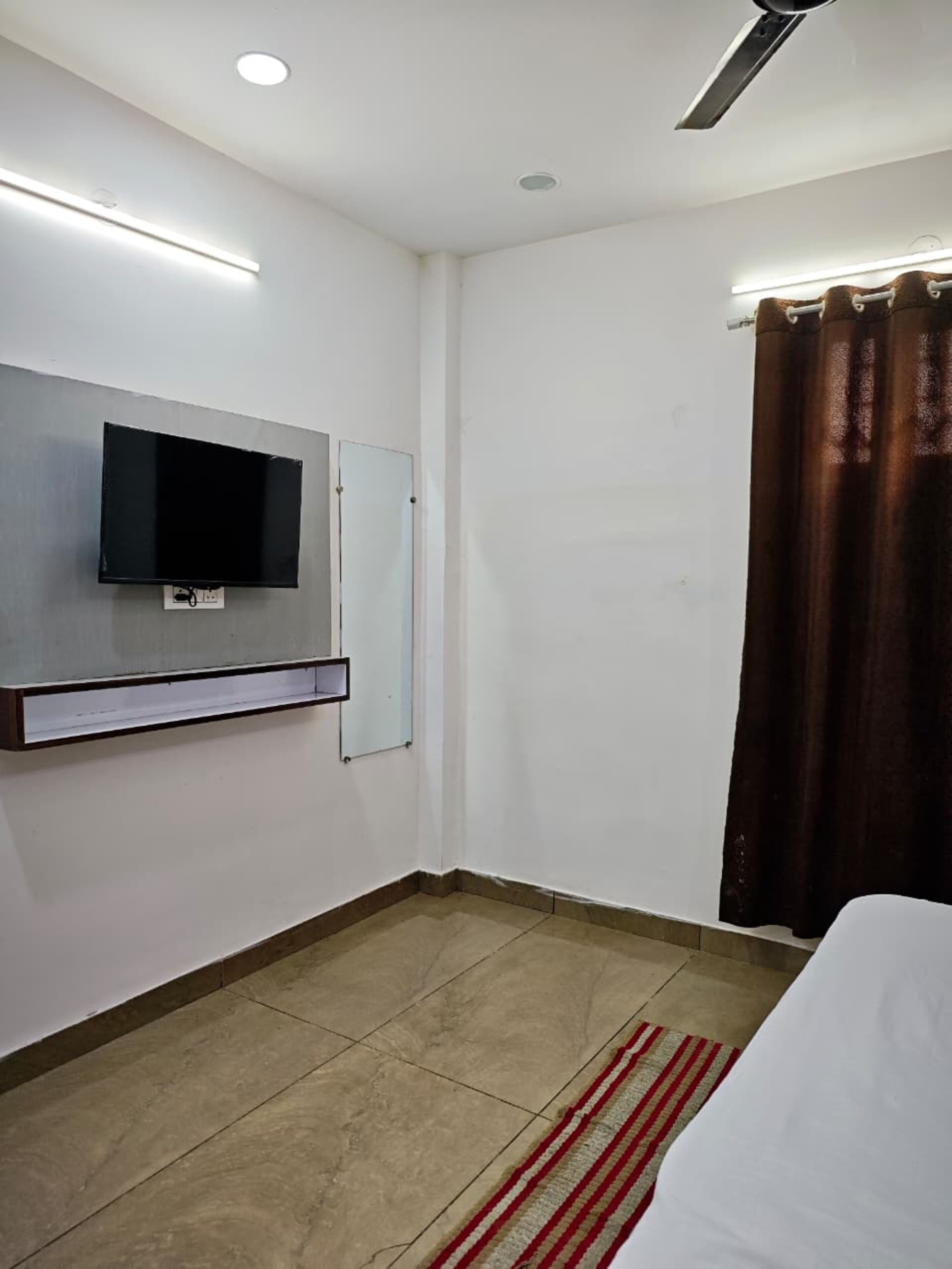 Habitación de hotel con televisor de pantalla plana montado en la pared, cama con ropa blanca, alfombra a rayas rojas y blancas, y cortina marrón.