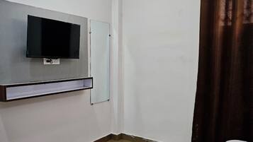 Habitación de hotel con televisor de pantalla plana montado en la pared, cama con ropa blanca, alfombra a rayas rojas y blancas, y cortina marrón.