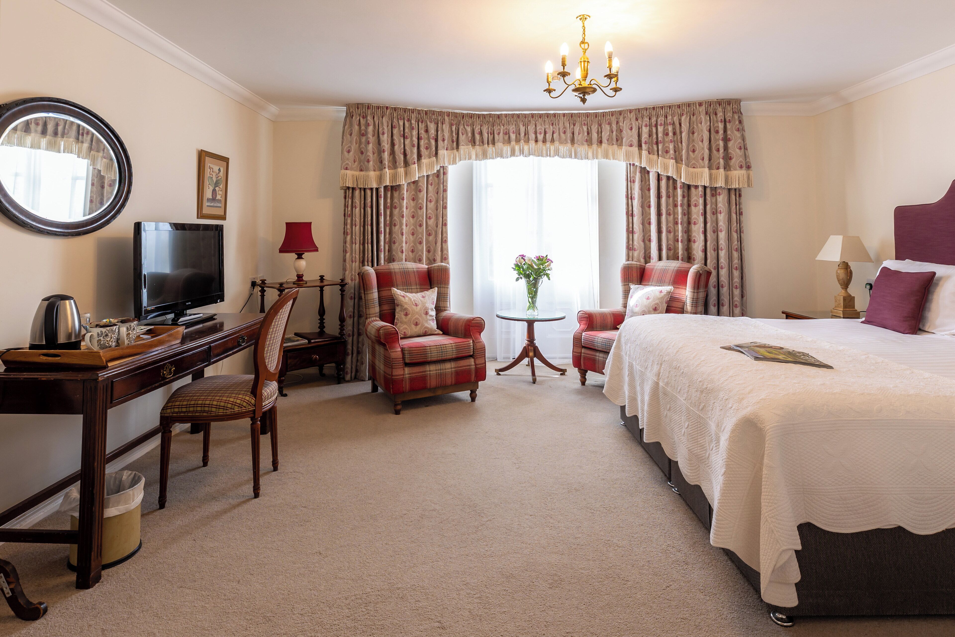 A hotel room with a bed, two armchairs, a desk with a television, and a small table with a vase of flowers.