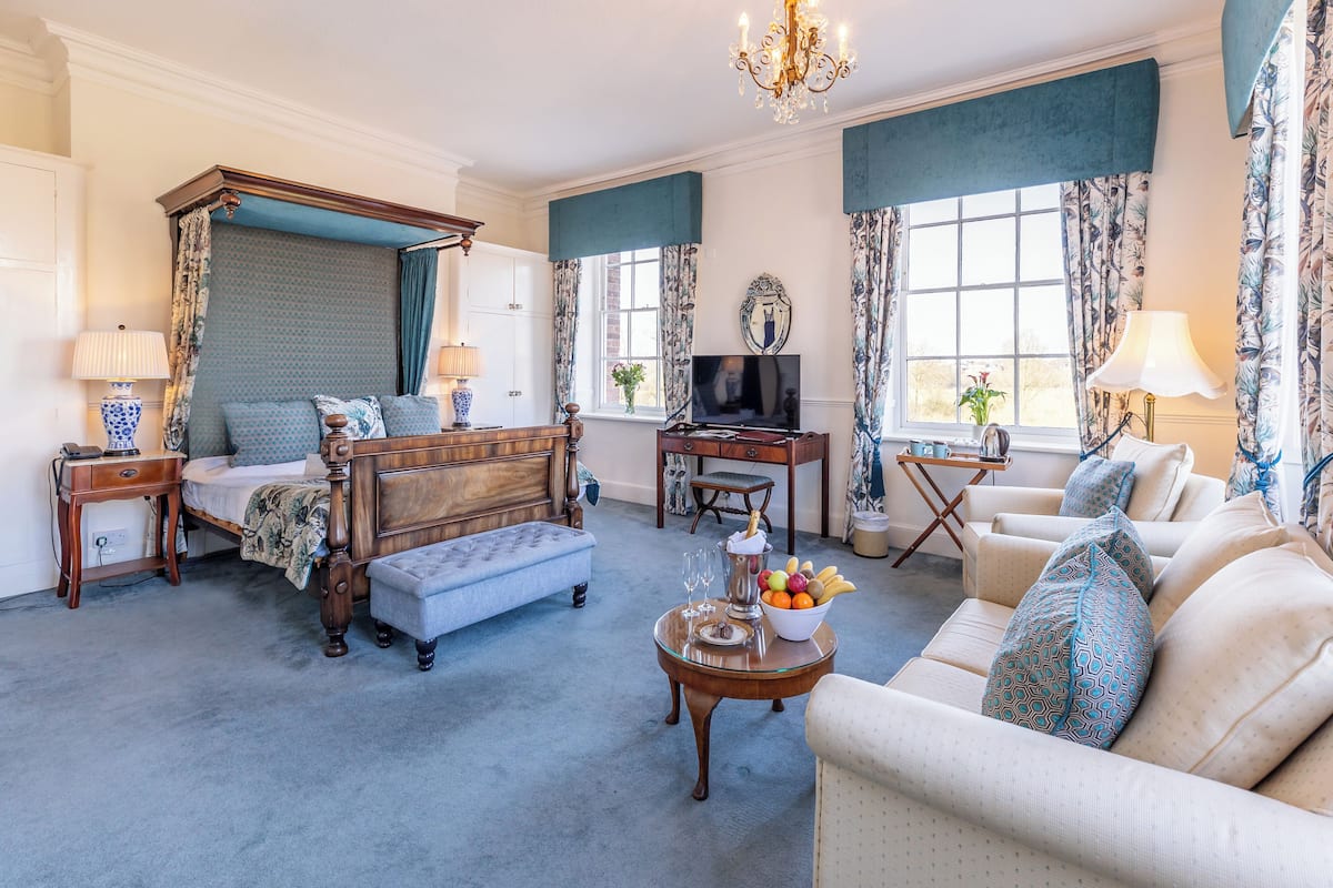 A well-appointed bedroom with a four-poster bed, a sitting area with a sofa and armchairs, a small dining table with a fruit bowl, and a television.