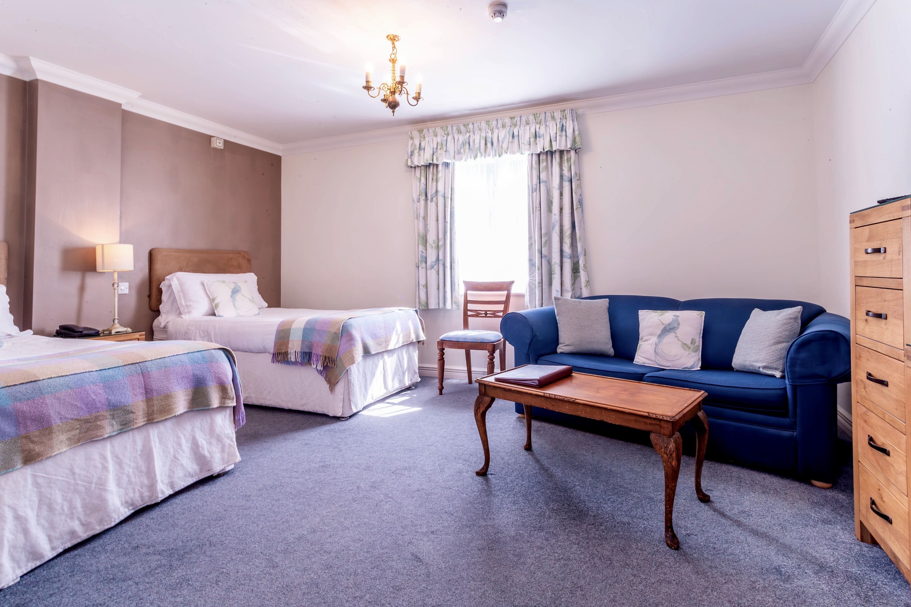 Une chambre d’hôtel avec deux lits, un canapé bleu, une commode en bois, une table basse en bois, une chaise et une fenêtre avec des rideaux.