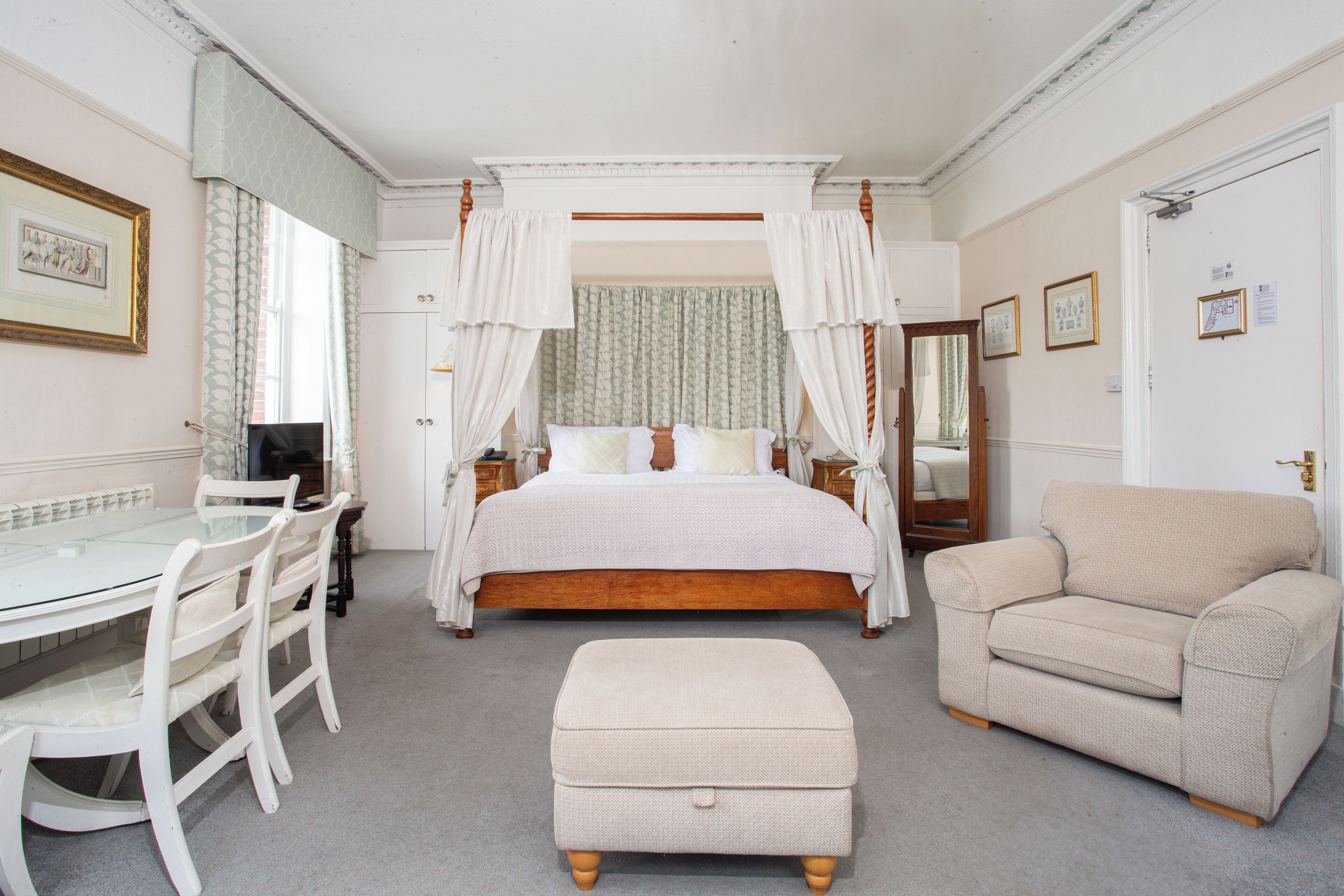 Ein Schlafzimmer mit Himmelbett, einer Sitzecke inklusive Sofa und Hocker, einem Esstisch mit Stühlen und einem Spiegel.
