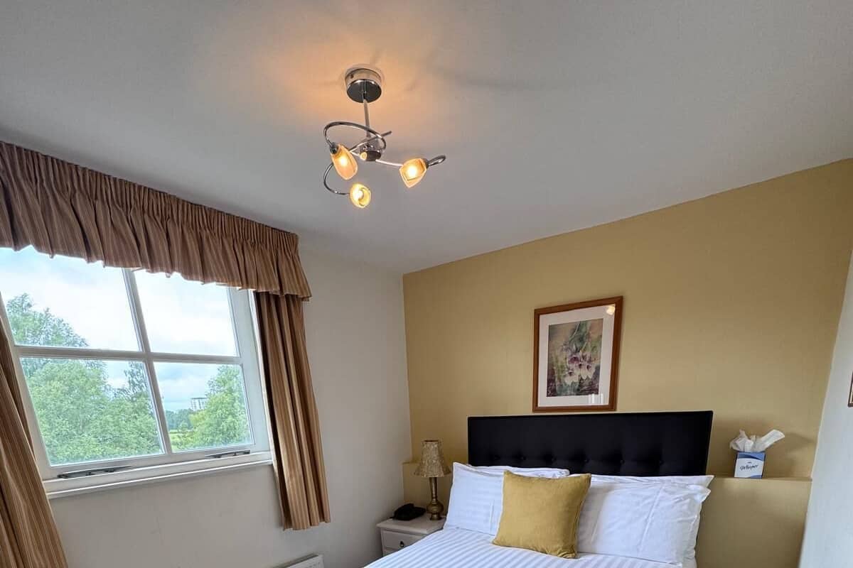 A bedroom with a bed, a nightstand, a lamp, a window with curtains, a radiator, and a ceiling light fixture.