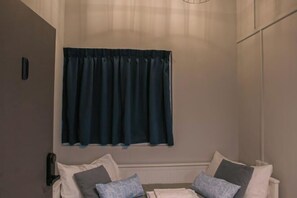 Una habitación con cama, ventana con cortinas oscuras y una lámpara fijada a la pared.