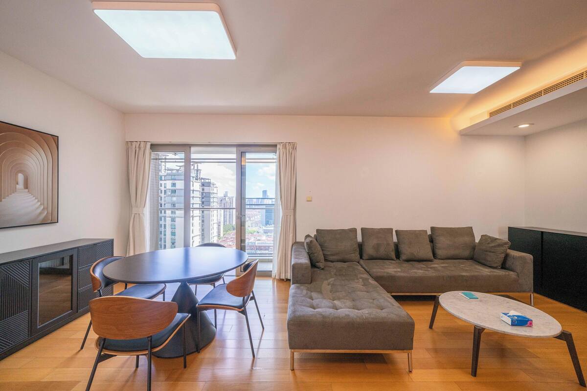 A modern living room with a round dining table, two sofas, and a large window with a city view.