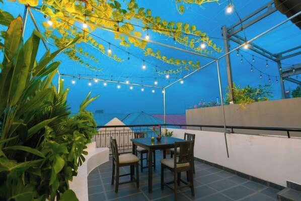 Un espacio al aire libre con patio, una mesa, sillas, luces colgantes y plantas en macetas.