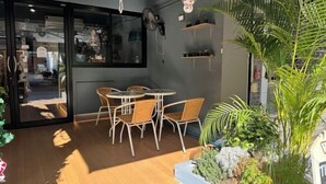 An indoor seating area with wicker chairs and a round table, a large potted plant, and a wall-mounted fan. - The Moment hostel (Bangkok)