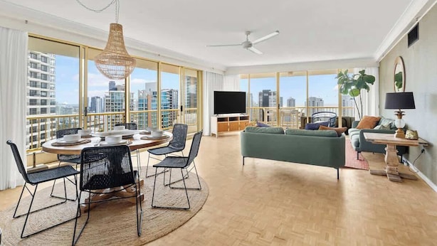 A spacious living room with a dining area, large windows with a city view, a flat-screen TV, and a ceiling fan. - Main Beach 3-Bedroom Apartment with Ocean Views (Main Beach)