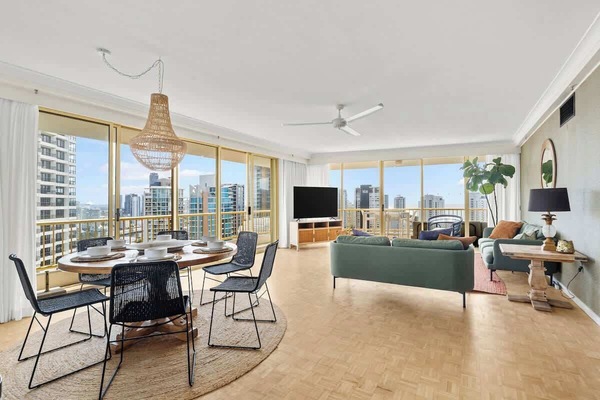 Take in the open-concept living and dining area featuring parquet flooring, a round dining setup with a woven pendant lamp, green sofas, and floor-to-ceiling glass doors overlooking high-rise city views.