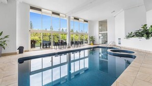 An indoor swimming pool with clear blue water, large windows, and a seating area with chairs and tables. - Main Beach 3-Bedroom Apartment with Ocean Views (Main Beach)