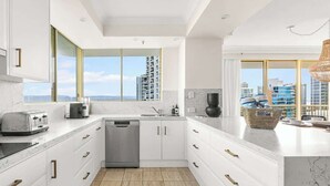 A modern kitchen with white cabinetry, a dishwasher, and a large window offering a city view. - Main Beach 3-Bedroom Apartment with Ocean Views (Main Beach)