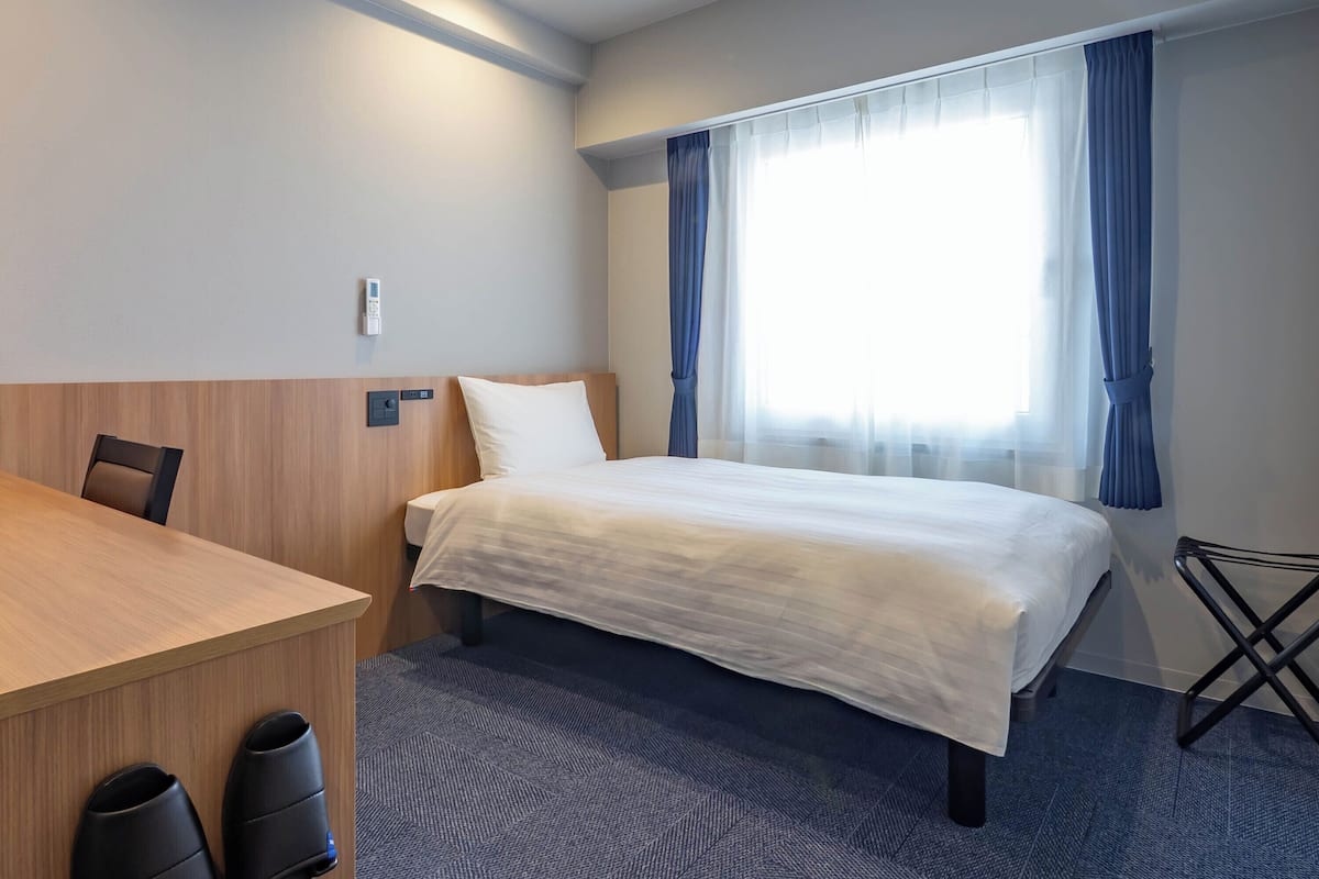 A hotel room with a bed, a desk, a chair, a shoe rack, and a window with curtains.