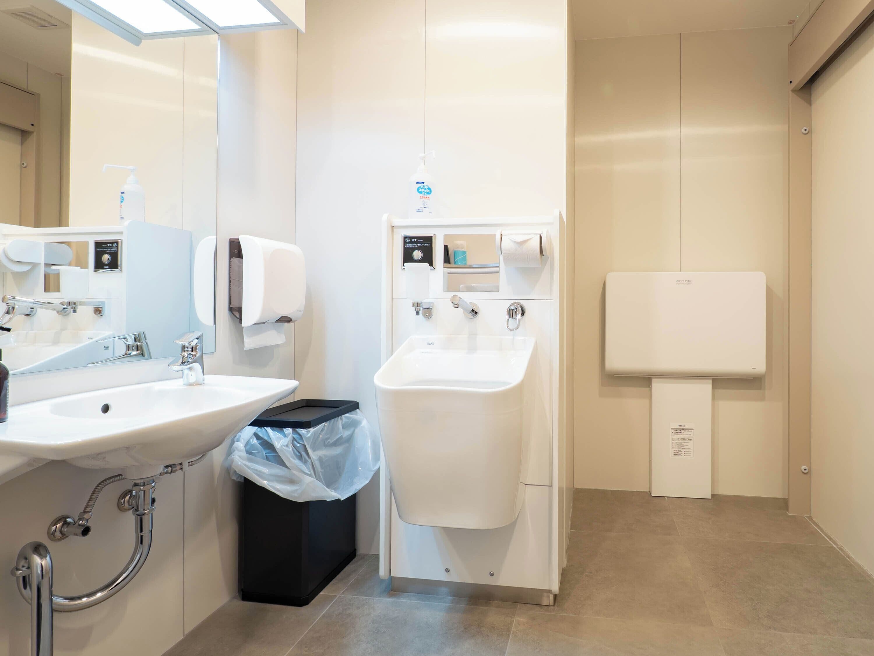 Un baño moderno con lavamanos, espejo, secamanos y dispensador de papel higiénico.