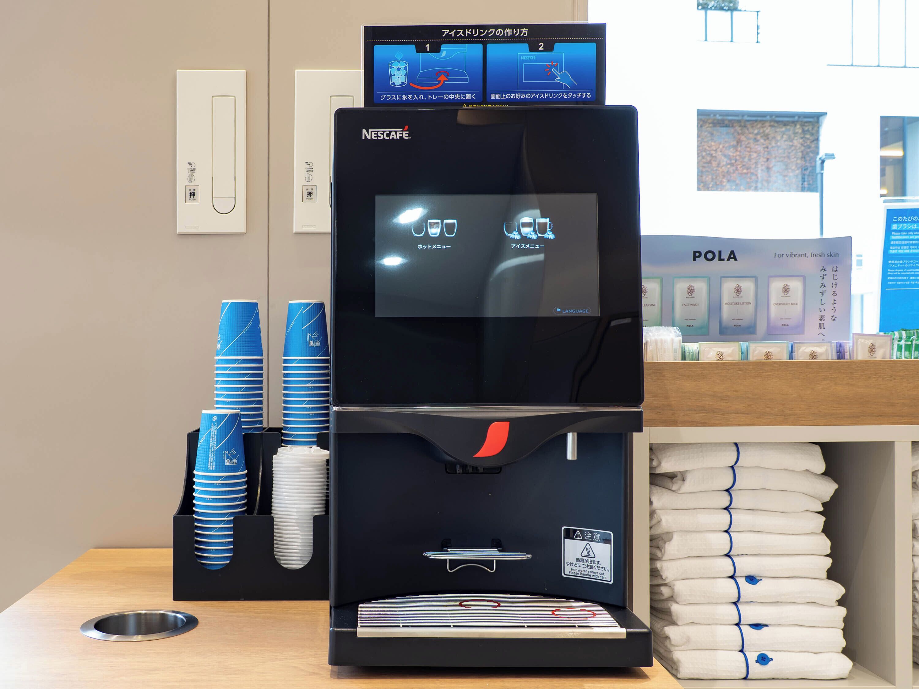 Een Nescafé koffieapparaat met een digitaal scherm, een stapel blauwe en witte bekers, en een plank met opgevouwen handdoeken.