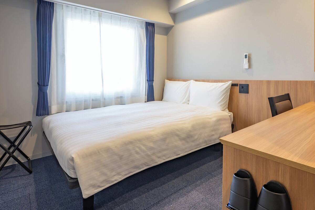 A hotel room with a bed, a desk, a chair, and a trash bin.