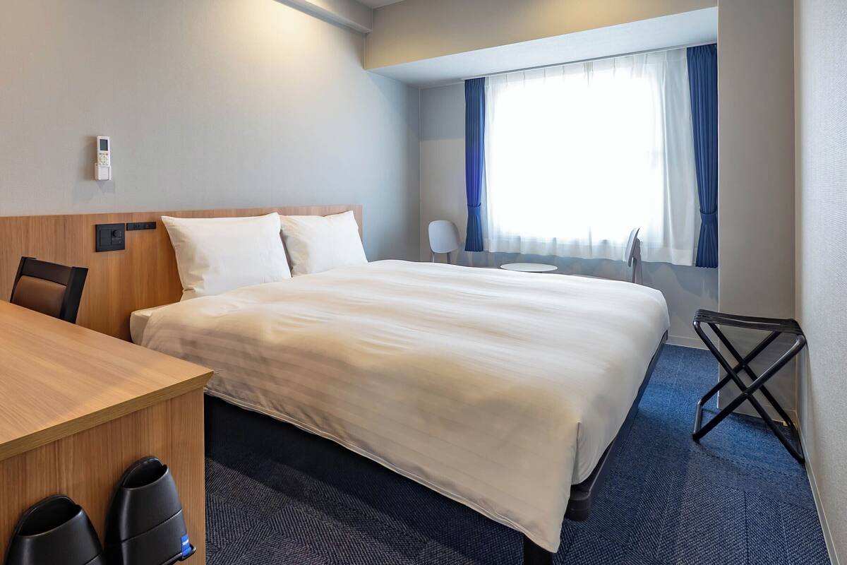 A hotel room with a bed, a desk, a chair, a small table, and a window with curtains.