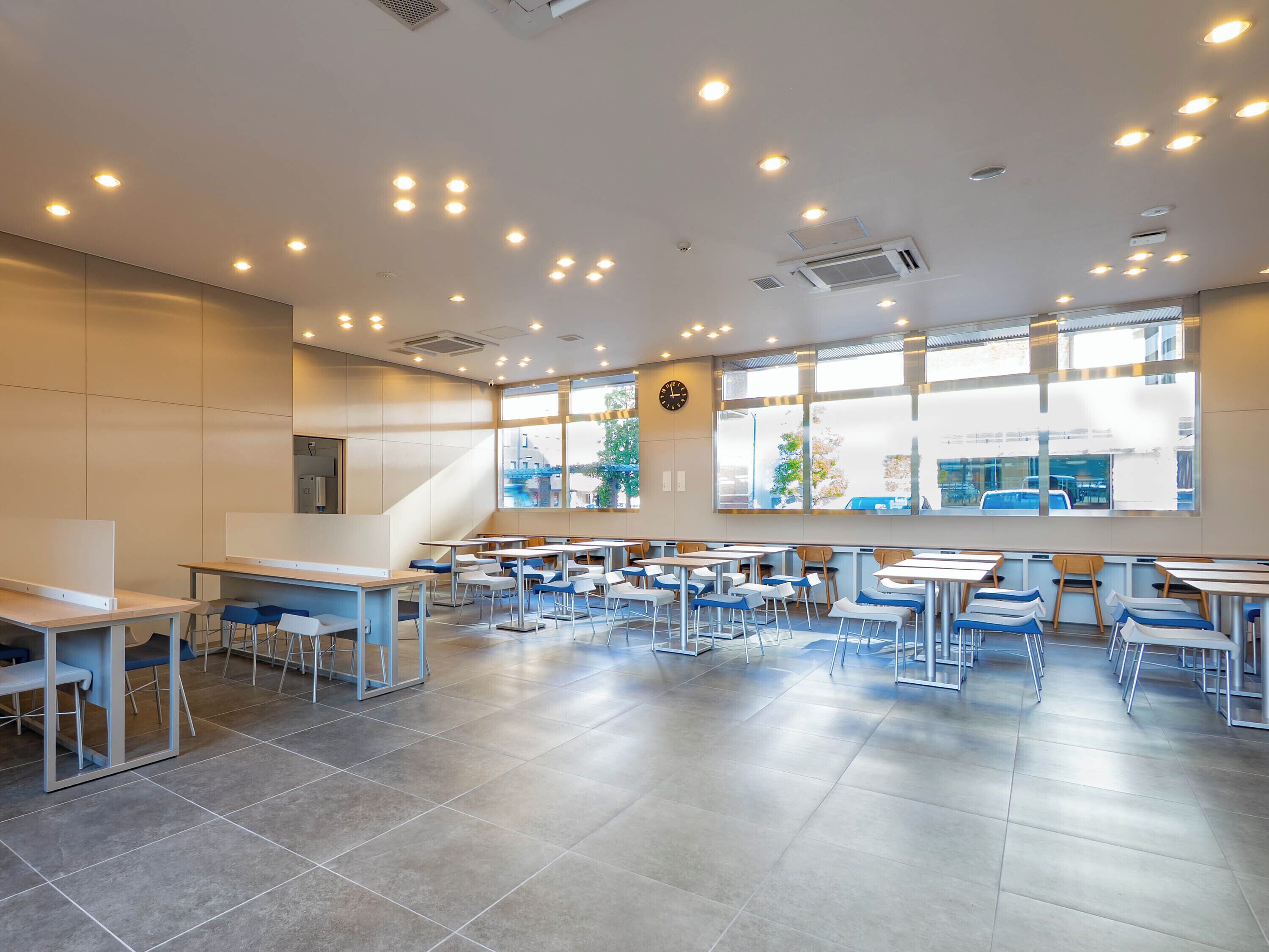 A spacious indoor area with multiple tables and chairs, large windows, and a clock on the wall.
