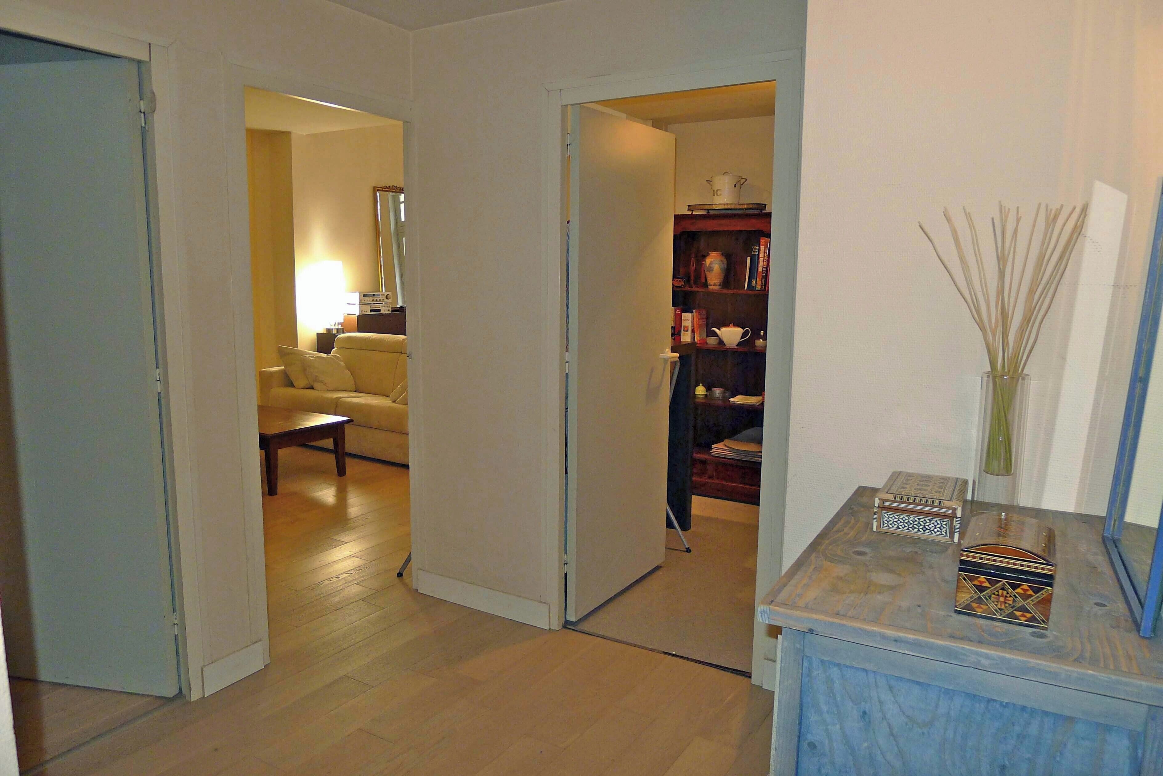 Una habitación con suelo de madera, una puerta blanca, un sofá, una mesa de centro y una estantería con diversos objetos.
