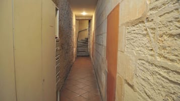 Un couloir étroit au sol carrelé, aux murs en pierre, et avec un escalier au fond.