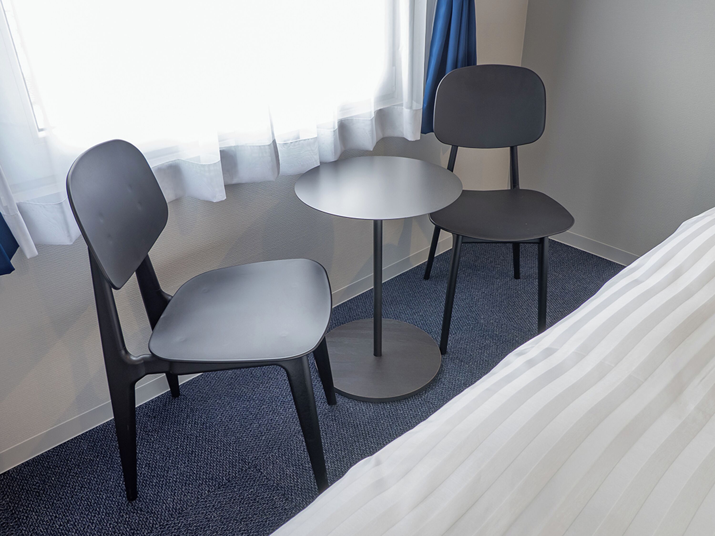 A hotel room with a bed, two chairs, a small table, and a window with curtains.