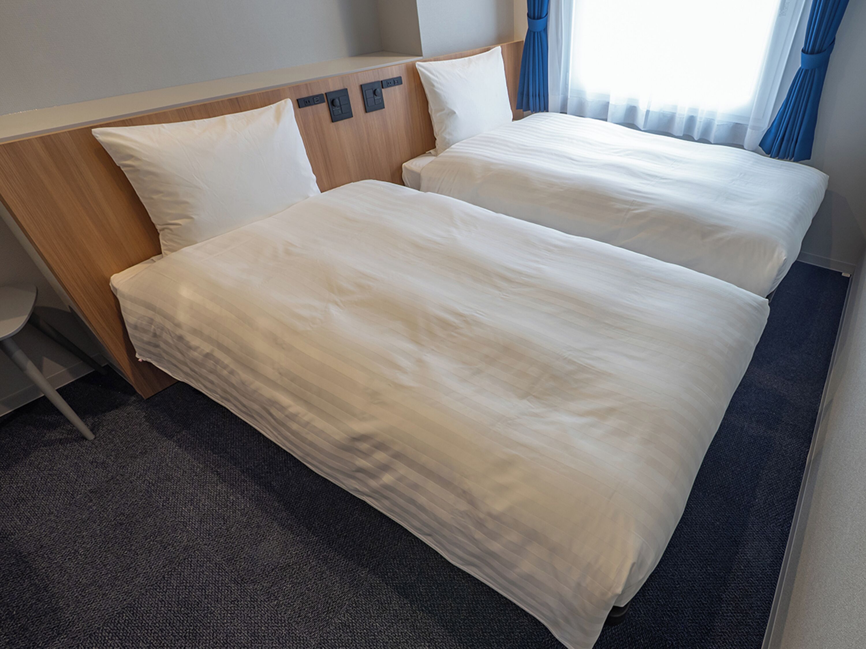 A hotel room with two beds, a wooden headboard, a small desk, and a window with curtains.