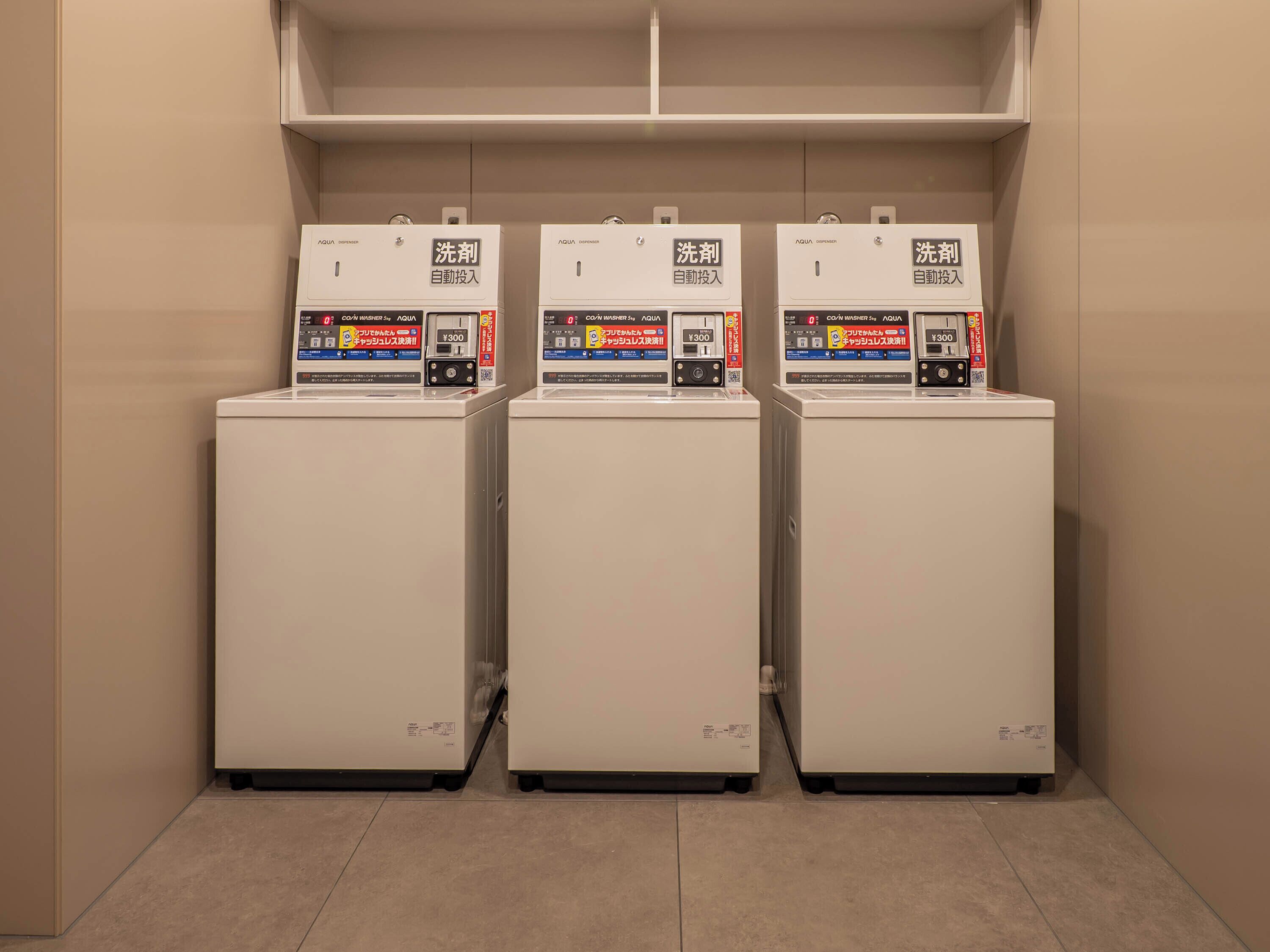 Three white laundry machines in a row with a shelf above them.