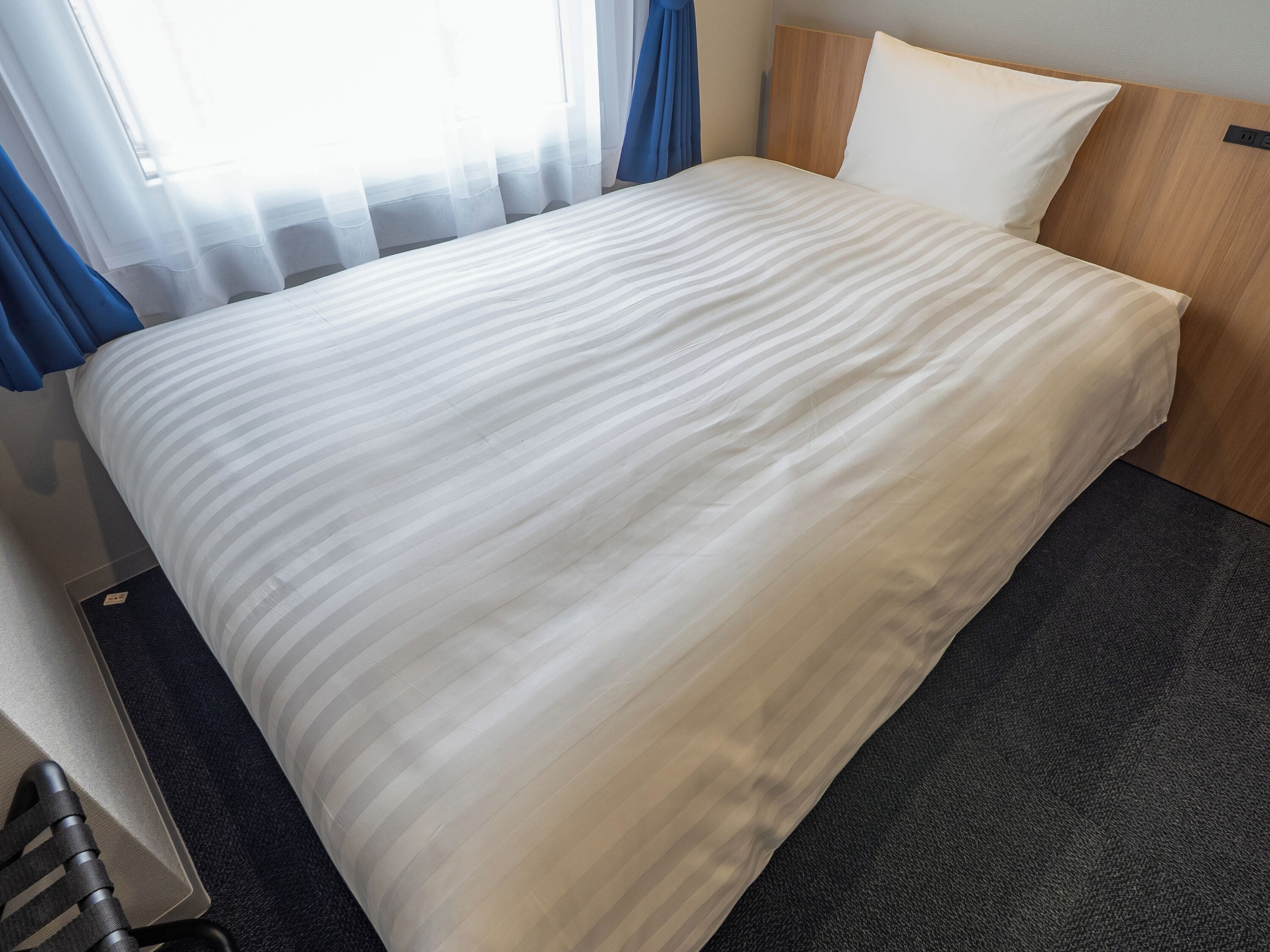 Una cama bien hecha con edredón blanco a rayas, cabecera de madera y suelo cubierto de moqueta oscura.