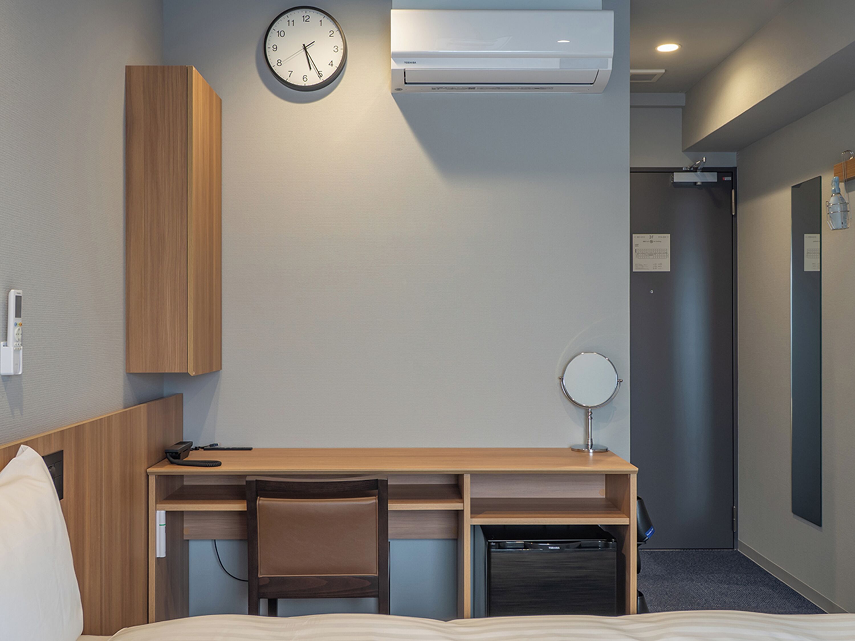 Habitación de hotel con escritorio de madera, un reloj, aire acondicionado, espejo y un refrigerador compacto.