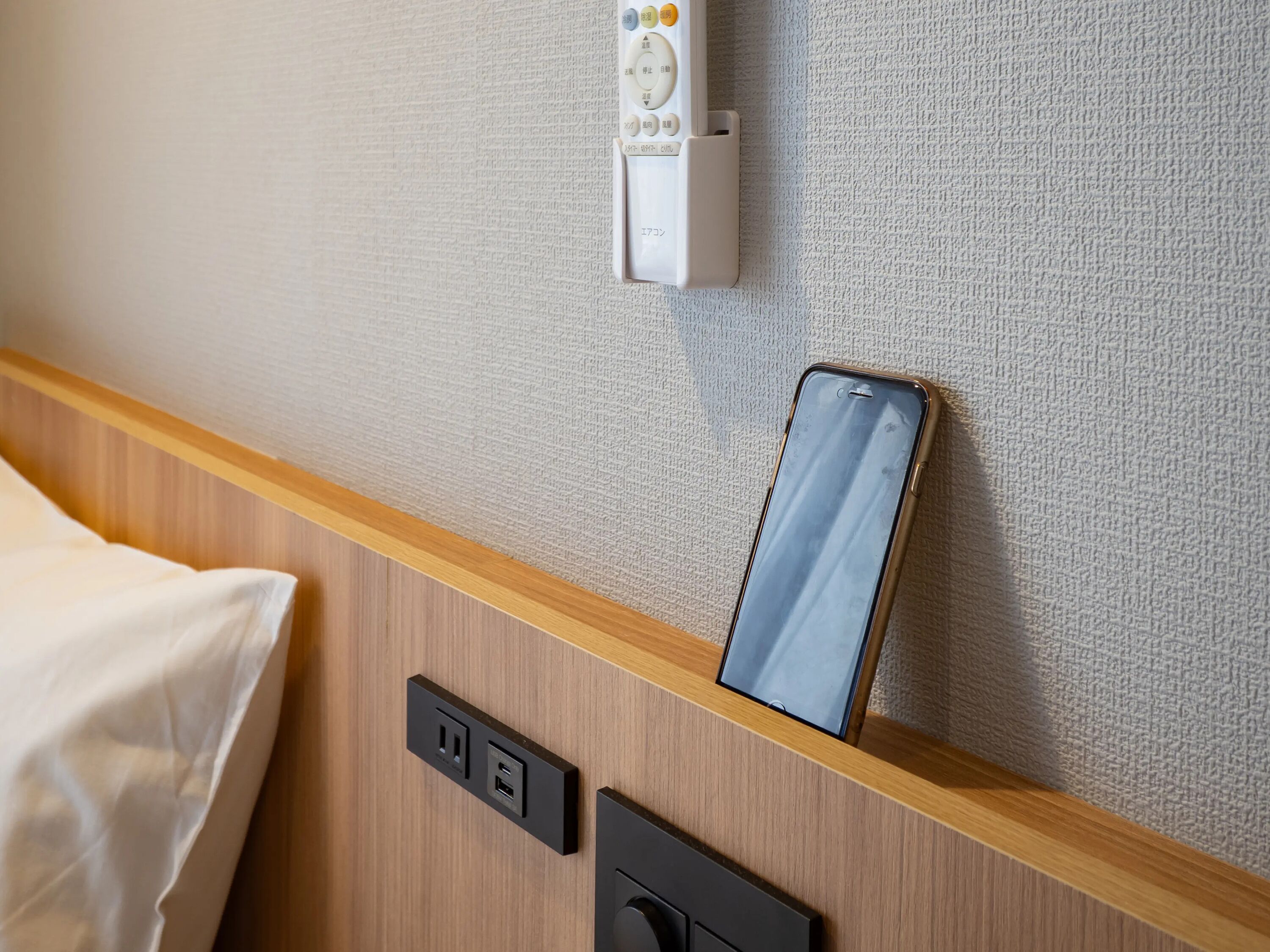 A hotel room with a wooden headboard, a bedside table with a smartphone, a power outlet, and a control panel on the wall.