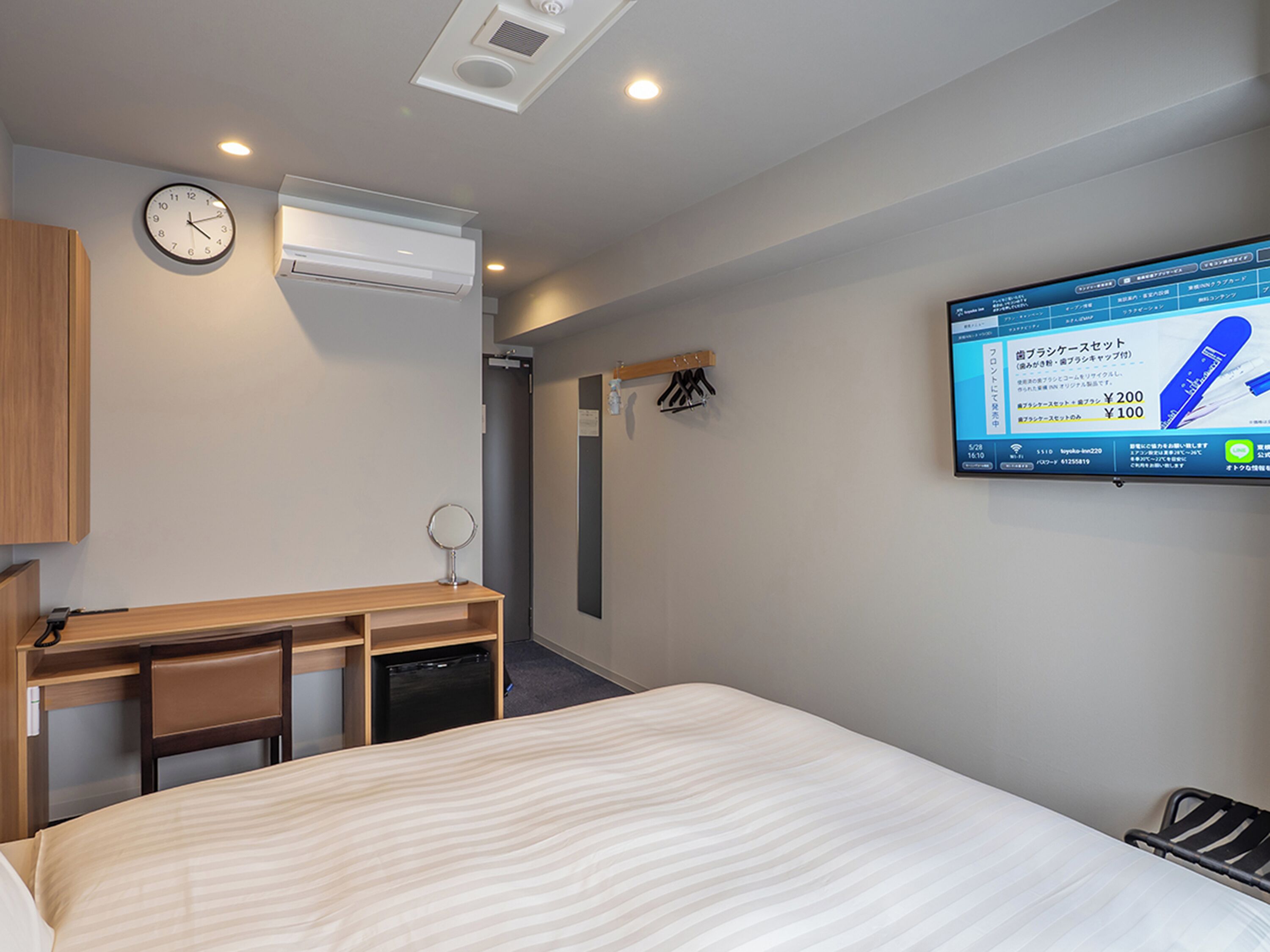 Hotelkamer met een bed, een bureau met stoel, een spiegel, een wandklok, een airconditioner en een televisie waarop een advertentie wordt weergegeven.