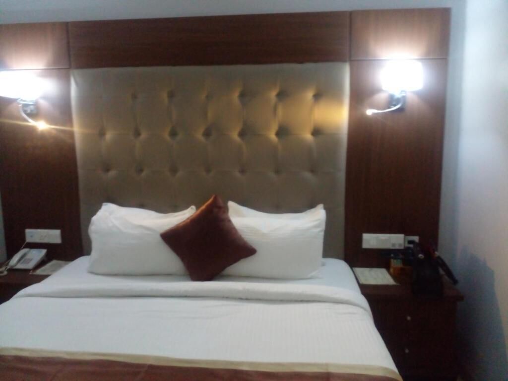 Een hotelkamer met een gestoffeerd hoofdeinde, een bed met witte lakens en een bruin kussen, plus nachtlampjes.