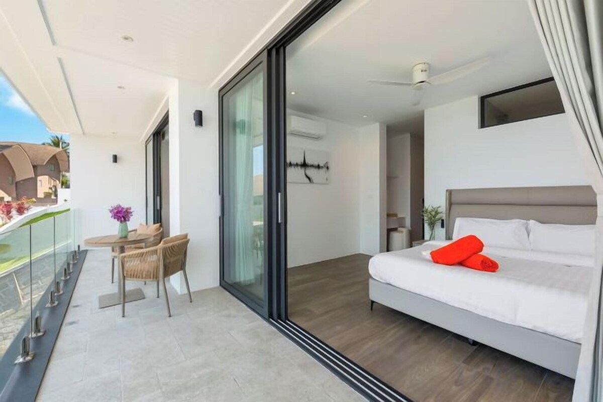Un dormitorio moderno con una cama grande, un balcón con mesa y sillas, y un ventanal corredero que da a un balcón.