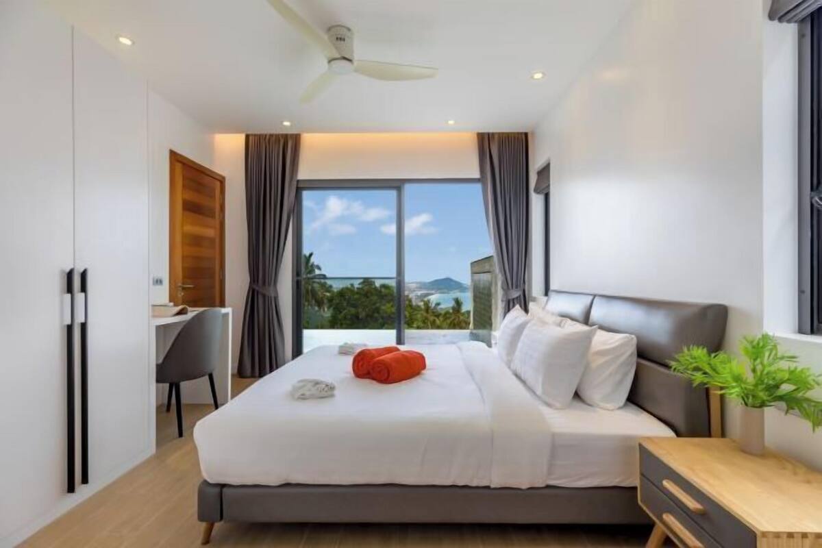 A modern bedroom with a large bed, a desk, and a view of the outdoors.