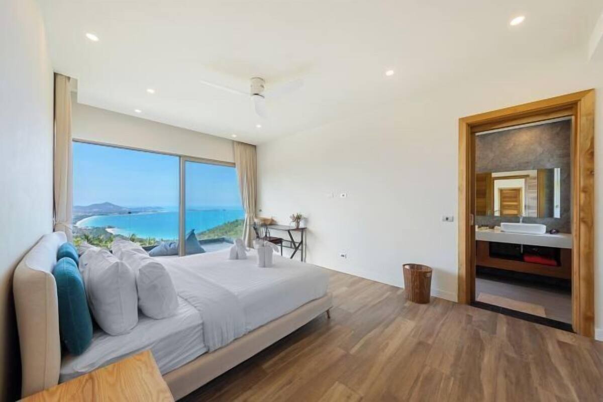 A bedroom with a large window, a bed, a desk, and a view of the ocean.