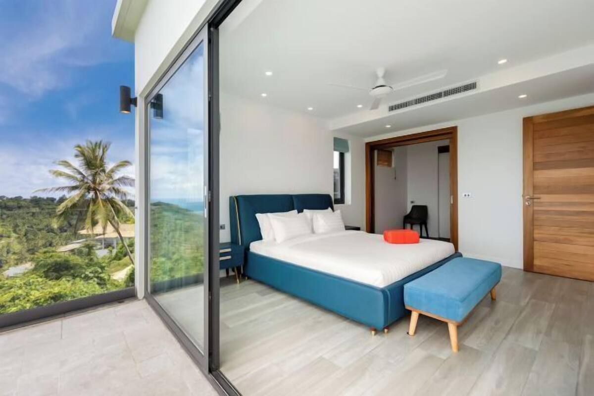 A modern bedroom with a large bed, a bench, and a sliding glass door leading to a balcony with a view of greenery.